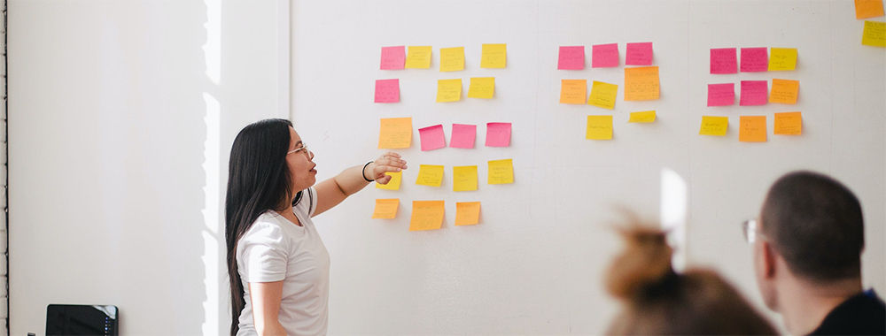 Photograph of woman standing in front of a whiteboard covered in sticky notes. She's presenting to a group of poeple around a table