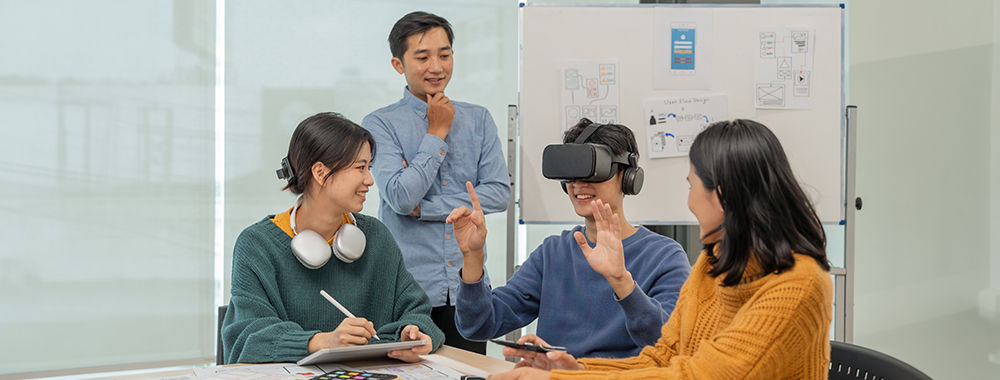 A group of people working together. One is standing, the other three are sitting around a desk. A whiteboard with sketches and notes on it is behind them. One of the people seated is wearing a VR headset. They are gesturing with their hands. The other thr