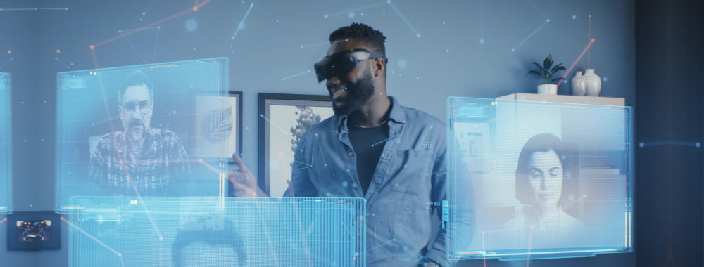 A man standing in a living room wearing a virtual reality headset. Surrounding him are several virtual screens, each showing a different person. They appear to be having a virtual meeting.