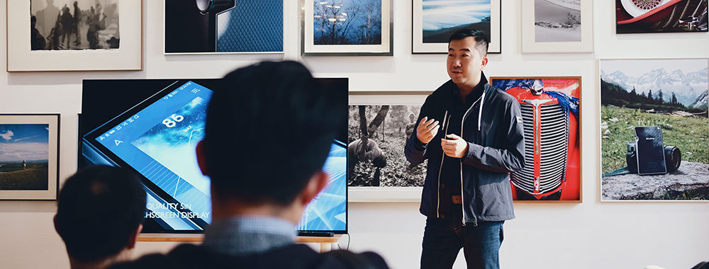 A man gives a talk in a photography gallery while standing next to a television screen displaying an image of a mobile phone.