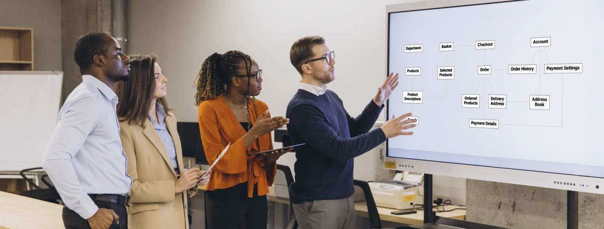 A UX design team standing together and reviewing an object-oriented UI architecture map displayed on a large interactive whiteboard screen.