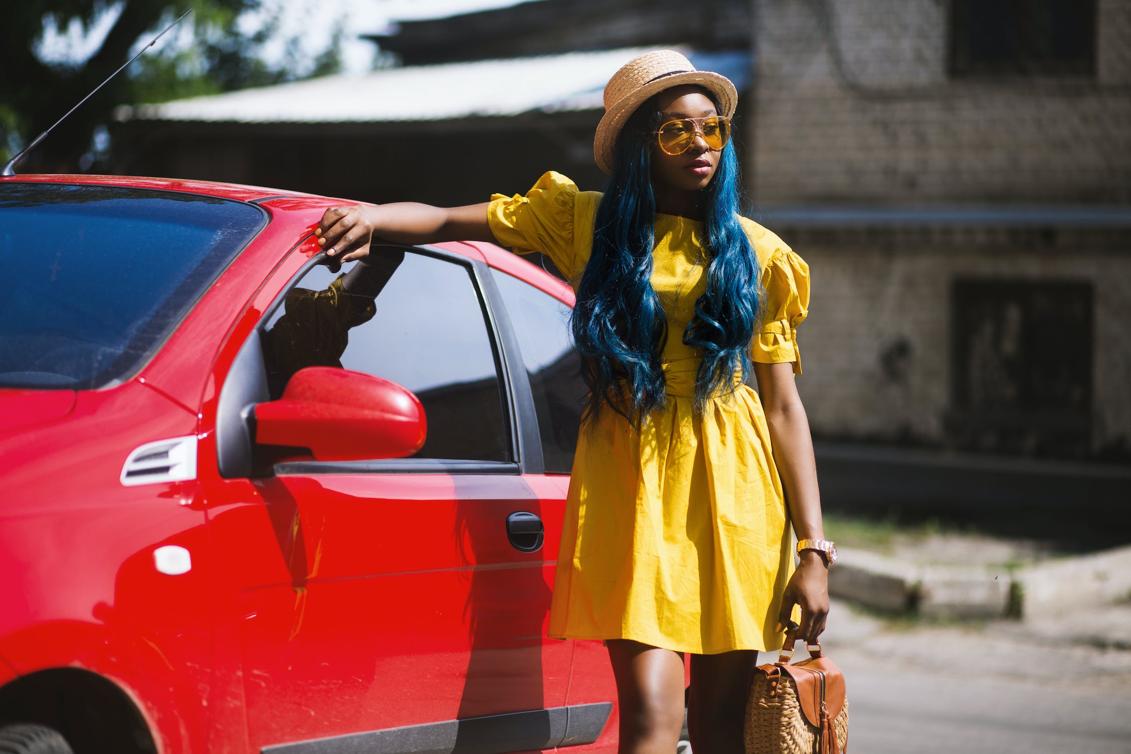 A vibrant image featuring a young woman wearing a sunny yellow dress with striking blue hair, set against the backdrop of a bold red car. 