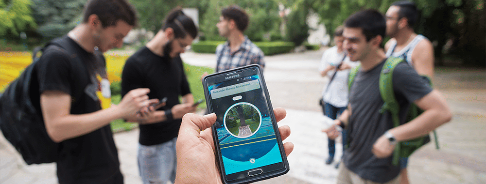 Photo of a person's hand holding a mobile phone. They're playing Pokémon GO. Around them are several other players. They look like they're in a park.