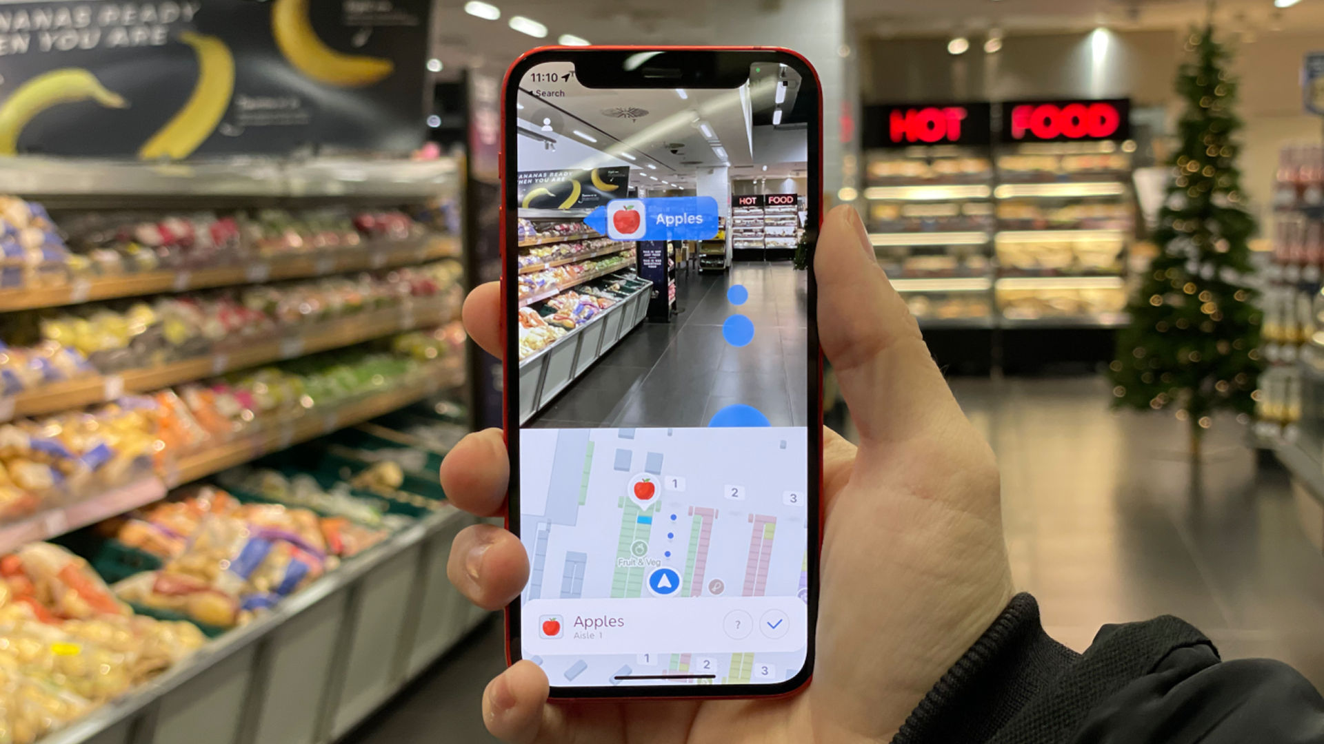 An image that shows how augmented reality works. A hand holds a mobile phone in front of a supermarket aisle. On the screen of the mobile phone is information that is digitally overlaid over the real environment, the supermarket.