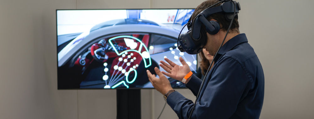 A man wearing a VR headset is looking at his hands. In front of him is a screen which shows what he's seeing inside the headset.