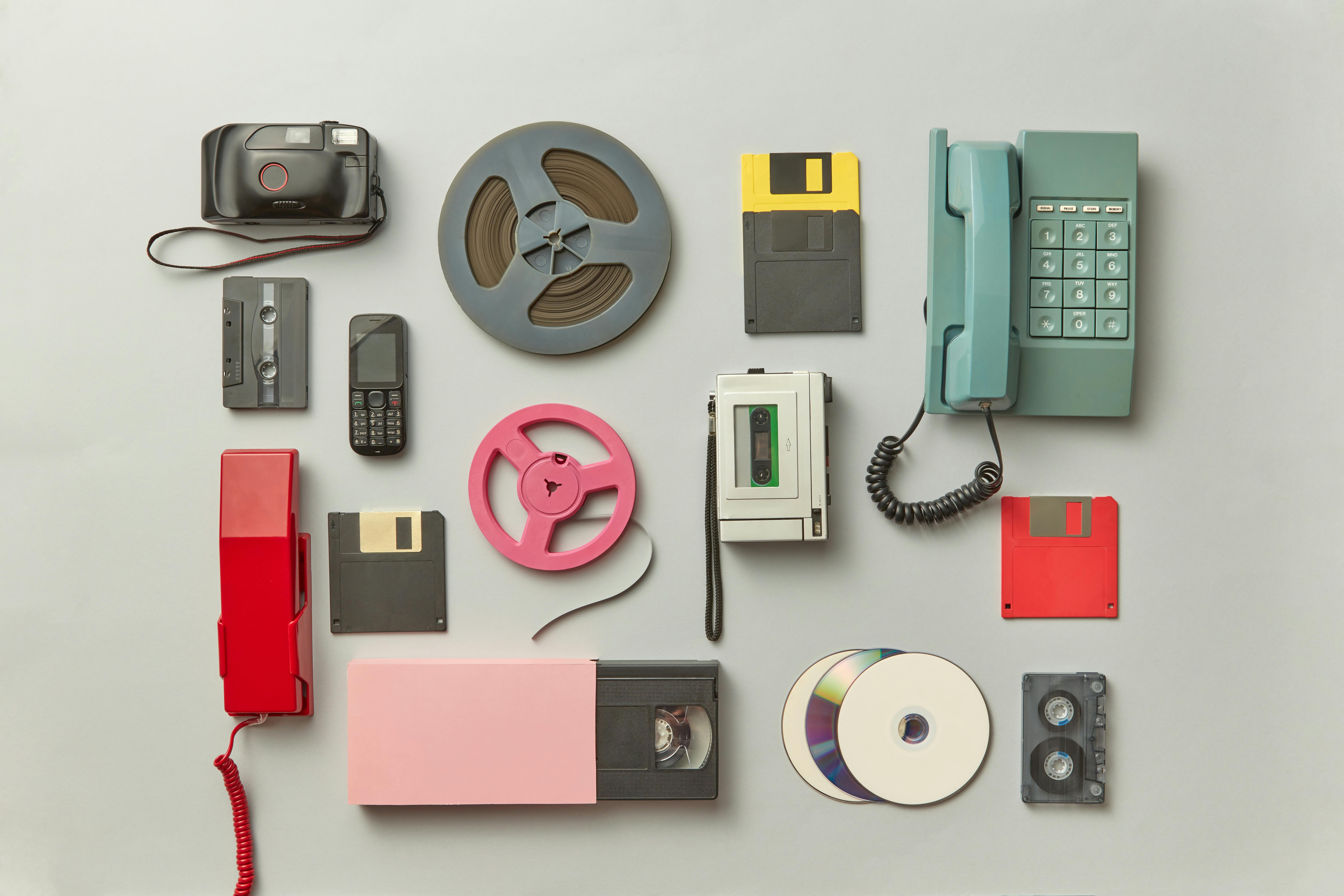 Flat-lay image of retro technology devices and storage media, including film reels, floppy disks, cassette tapes, a VHS tape, CDs, a point-and-shoot camera, an old mobile phone, and corded telephones arranged on a neutral background, illustrating vintage electronics and obsolete formats.