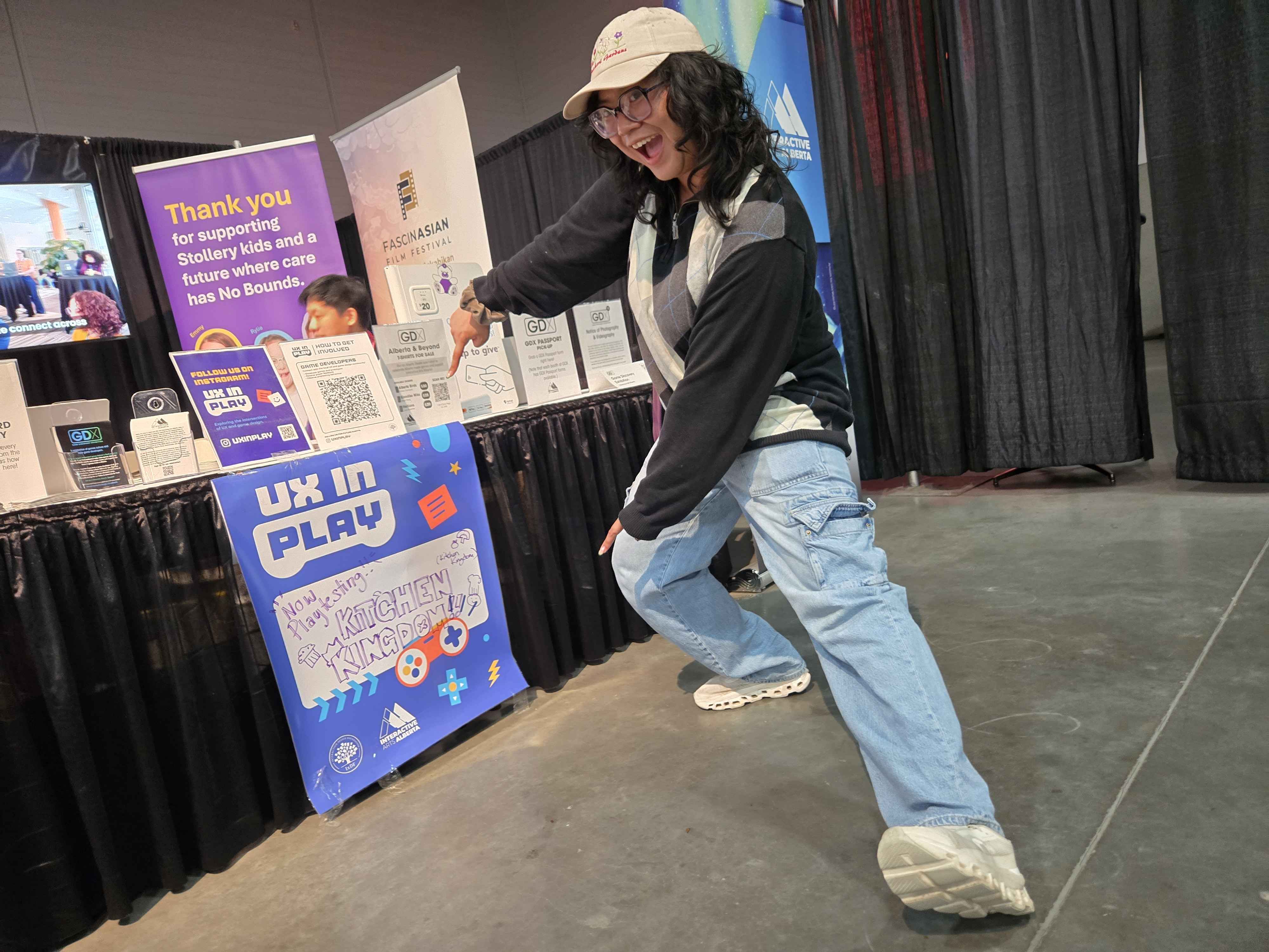 IxDF Edmonton volunteer pointing to our playtesting signage at the IAA booth. The sign reads,